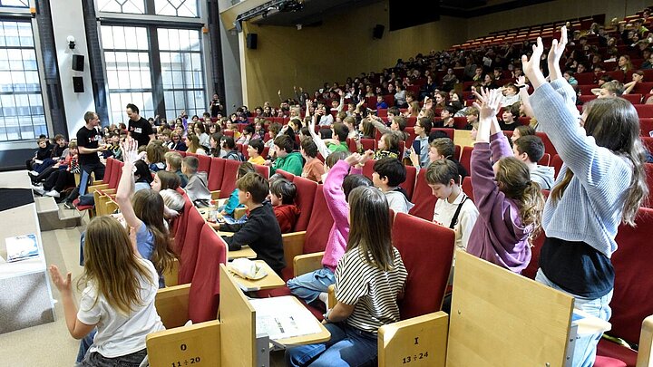 Sala wykładowa wypełniona widownią, duża część zgromadzonej młodzieży ma wyciągnięte do góry ręce, zgłaszając się do odpowiedzi.